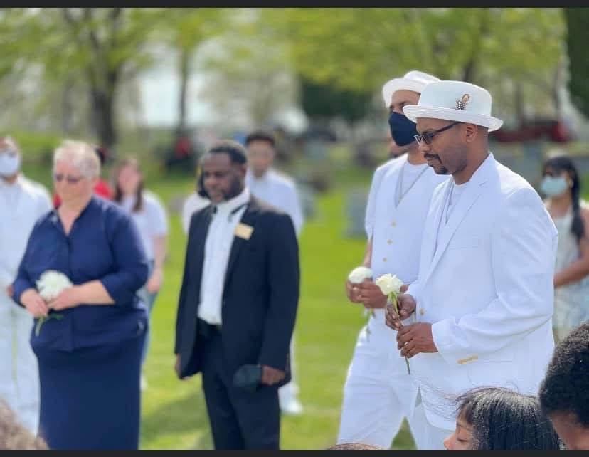 Mourners in white suits holding white roses at the graveside